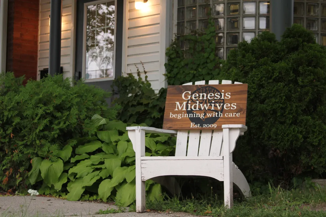 A sign which reads Genesis Midwives sitting on a Muskoka chair outside of the clinic,